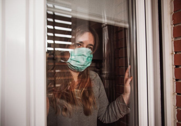 A young woman in quarantine wearing a mask and looking through the window/divulgadoras-crisis-coronavirus