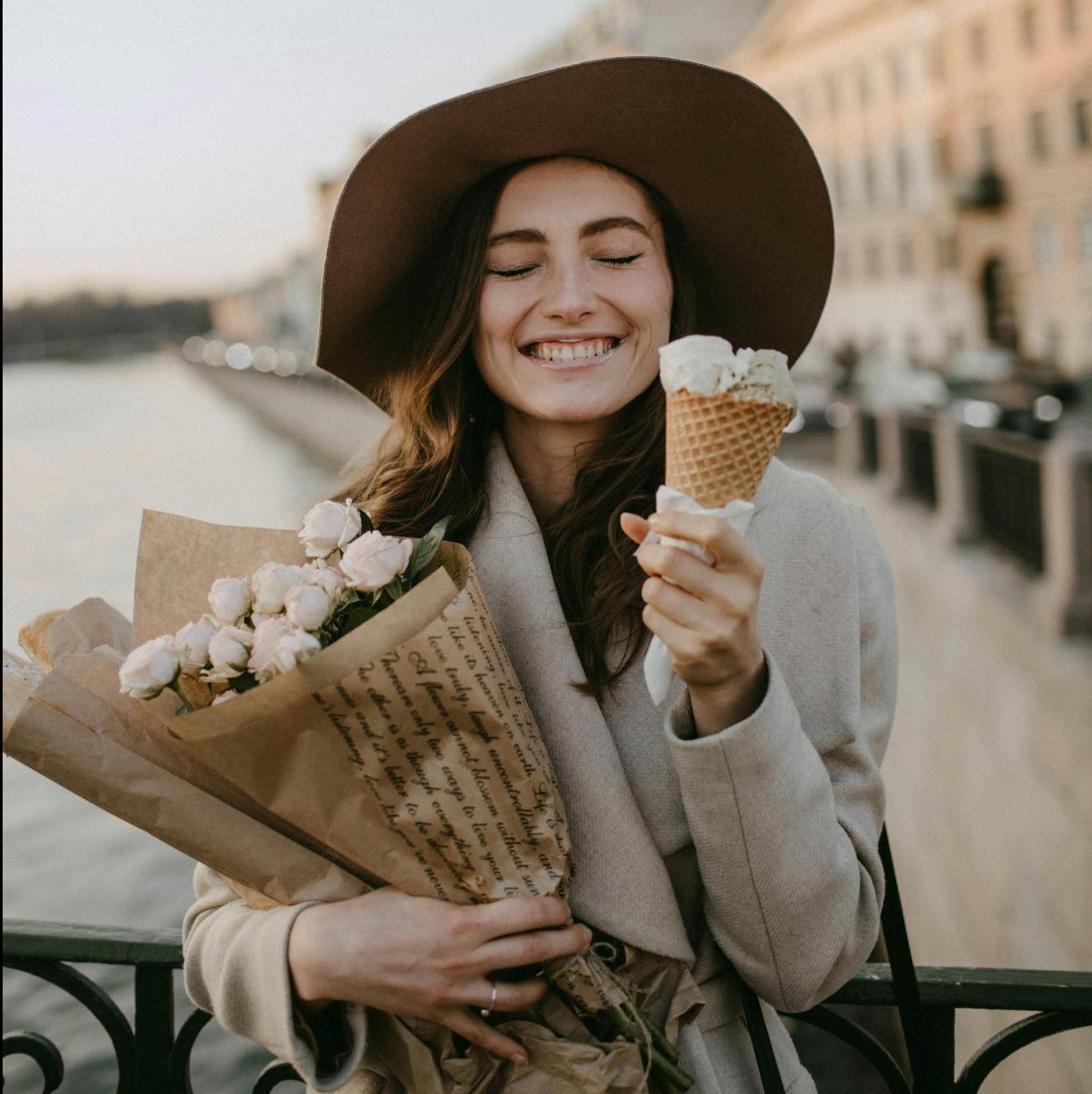 Mujer feliz tomando helado y sosteniendo flores. /PEXELS